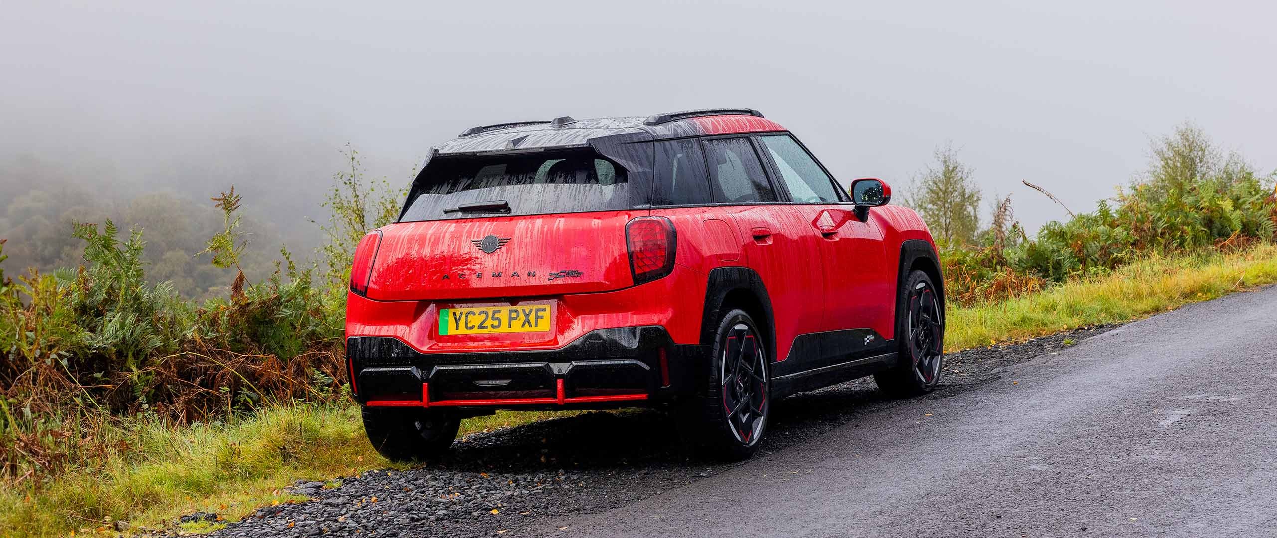 Rear side view of parked all-electric MINI Aceman in British Racing Green in car park with all doors open and coloured wall in the background